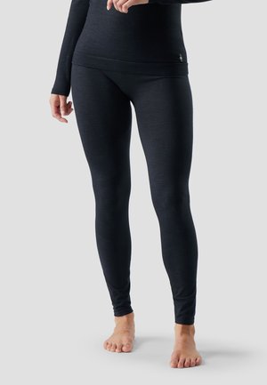 Person wearing black fitted leggings and long-sleeve top, standing barefoot against a plain background.