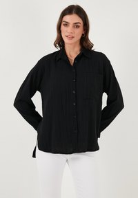 Black button-up shirt with a textured fabric, long sleeves, a chest pocket, and side slits; pairs well with white pants.