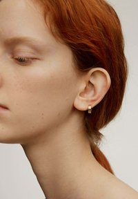 Pendiente de aro dorado con un diseño texturizado, sobre un tono de piel suave y con un cabello rojo contrastante. La vista de perfil resalta el pendiente.
