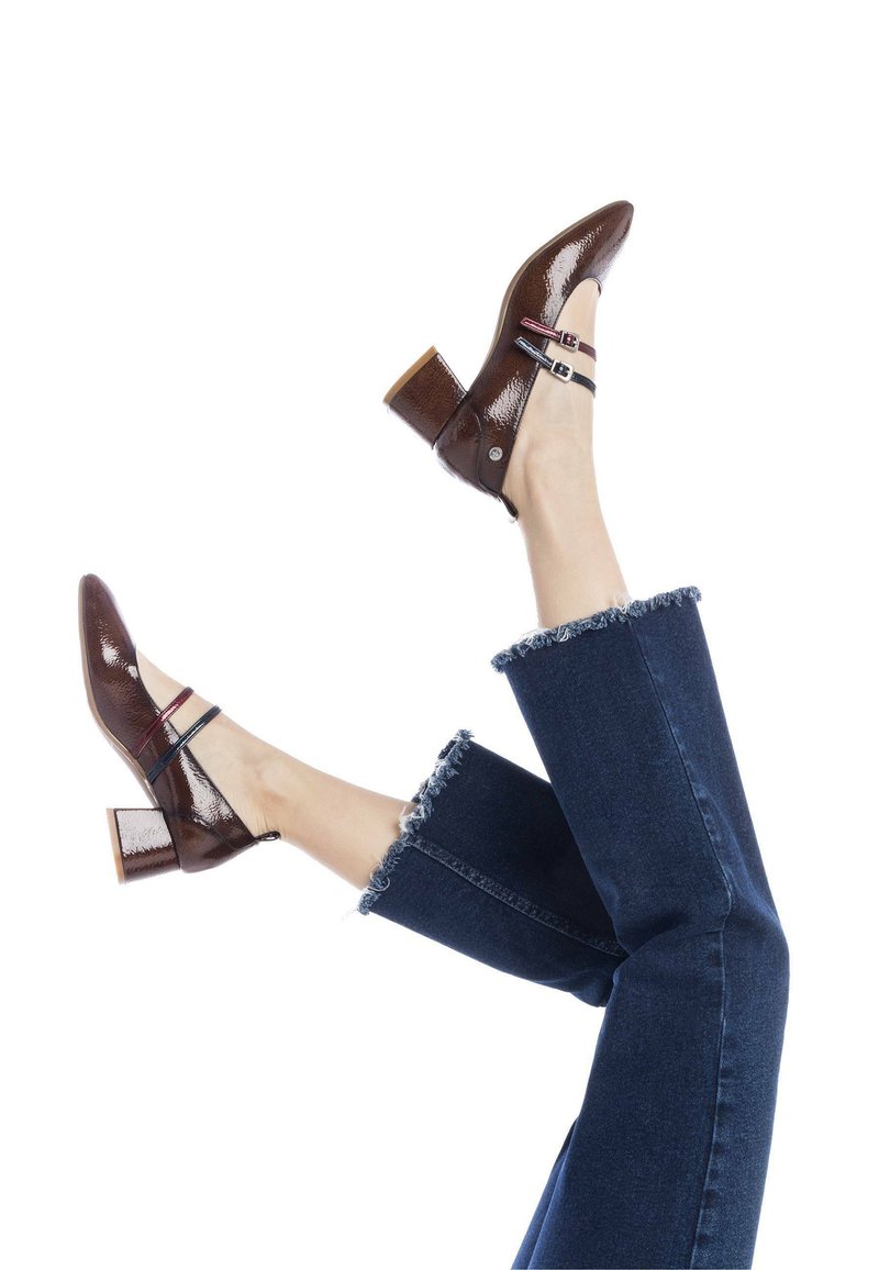 Tacones de charol marrón con un tacón grueso, que presentan tiras negras y rojas a lo largo del mediopié. El modelo lleva puestos unos pantalones vaqueros oscuros deshilachados.