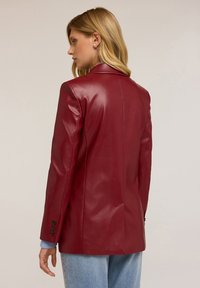 Blazer en cuir rouge avec une silhouette ajustée, des revers en pointe et des poignets à boutons. Texture lisse avec des détails de couture simples.