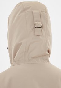 Vaaleanbeige huppu takki, jossa on korkea kaulus ja selässä oleva hihna sekä metallinen solkiyksityiskohta. Sileä ja kevyt kangas.