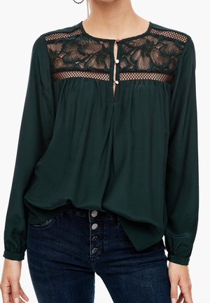 Blouse à manches longues vert foncé avec dentelle florale et empiècement en tulle, col en goutte d'eau boutonné, portée avec un jean bleu foncé à quatre boutons.