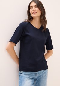 Námořnická modrá tričko s krátkým rukávem vyrobené z texturované látky, s kulatým výstřihem, v kombinaci se světle modrými džíny. Jednoduchý design, bez vzorů.