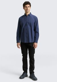 Camicia a maniche lunghe a scacchi blu navy con chiusura a bottoni, indossata con pantaloni neri e scarpe scure. Tessuto liscio con un design del colletto classico.