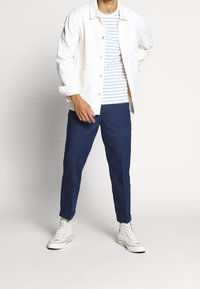 Valkoinen farkkutakki sinivalkoisen raidallisen paidan päällä, yhdistettynä tummansinisiin housuihin ja valkoisiin korkeavartisiin lenkkareihin.