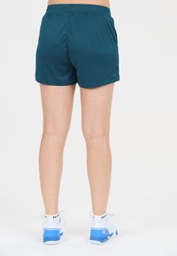 Person der står op, iført teal træningsshorts, hvide sokker og hvide og blå sneakers, vist fra ryggen mod en hvid baggrund.