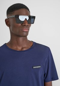 Occhiali da sole neri angolari con lenti riflettenti, indossati con una maglietta girocollo blu navy con un patch logo sul lato sinistro del petto.