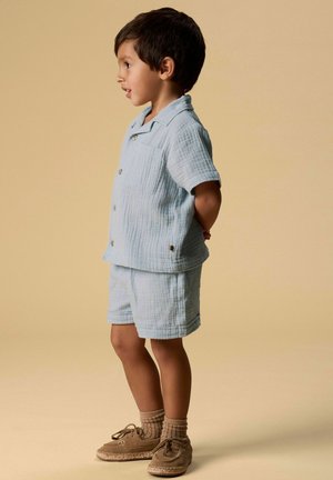 Jeune garçon debout de profil, portant une chemise bleu clair à manches courtes et un short, des chaussettes beige et des chaussures marron à lacets, sur un fond beige.