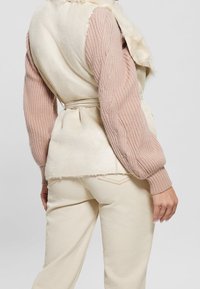 Donna che indossa pantaloni color crema, un gilet in finta pelliccia color crema con cintura e un maglione a coste rosa polveroso con maniche a sbuffo.