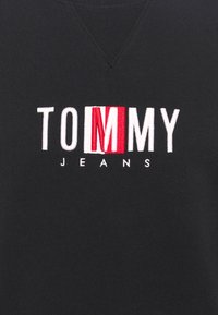 Tissu noir avec le texte blanc "TOMMY" et la lettre "M" mise en évidence en rouge, accompagné d'un texte plus petit en blanc "JEANS" en dessous.