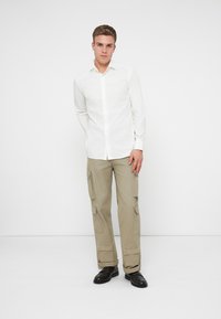 Hvit skjorte med knapper, beige cargo bukser med kne lommer, og svarte sko. Modellen står mot en enkel bakgrunn.