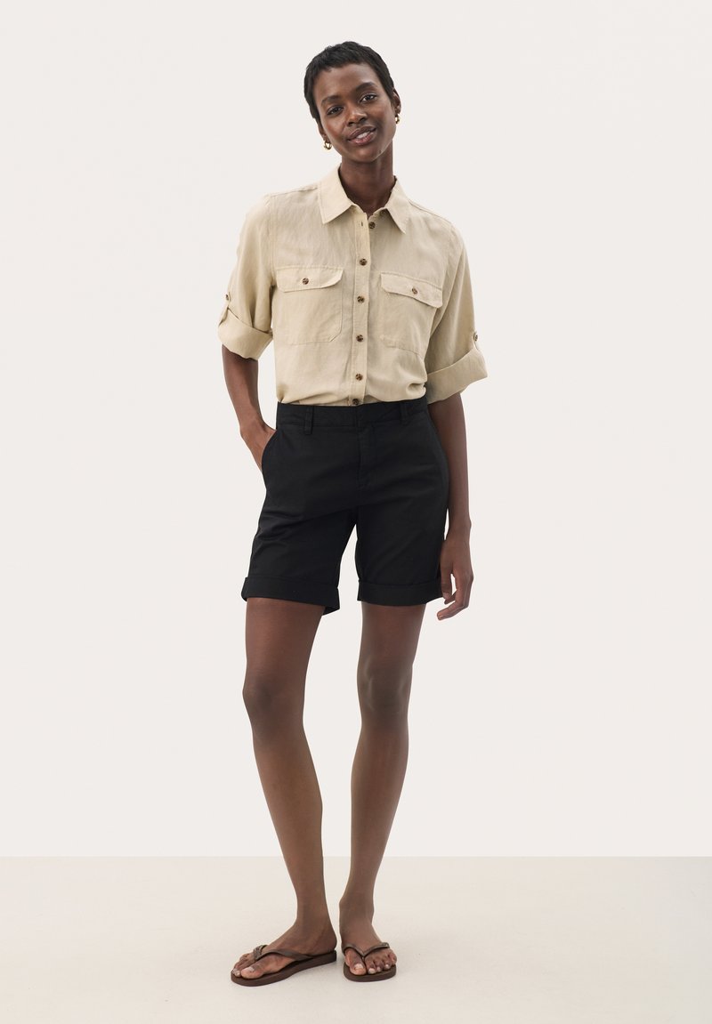 Chemise à manches courtes beige clair avec deux poches poitrine ; short noir retroussé ; tongs beiges ; arrière-plan neutre.