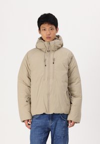 Beige gepolsterte Jacke mit hohem Kragen, zwei vorderen Reißverschlüssen und elastischen Bündchen. Sie hat eine glatte Oberfläche und Kordelzüge an der Kapuze. Blaue Jeans aus Denim.
