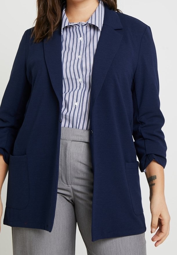 Femme portant un blazer bleu marine par-dessus une chemise à rayures bleues et blanches boutonnée et un pantalon taille haute gris clair.
