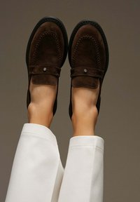 Brown suede loafers with a stitched design, square toe, and flat sole; metallic accent on the strap; styled with white trousers.