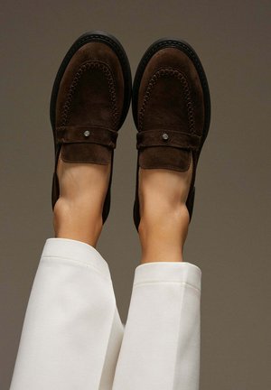 Brown suede loafers with a stitched design, square toe, and flat sole; metallic accent on the strap; styled with white trousers.