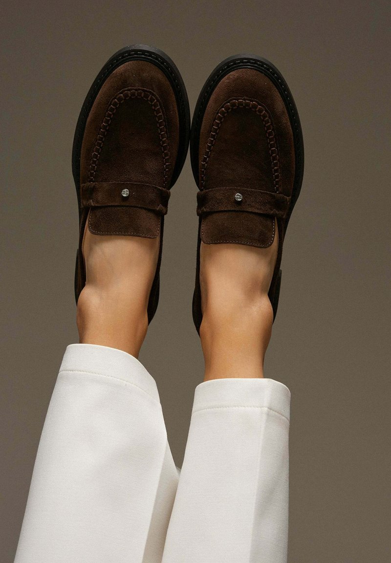 Brown suede loafers with a stitched design, square toe, and flat sole; metallic accent on the strap; styled with white trousers.