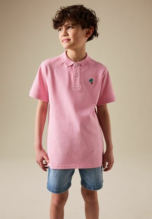 Băiat cu păr creț, purtând un tricou polo roz cu un mic emblema verde cu palmier și pantaloni scurți din denim, stând în fața unui fundal simplu.