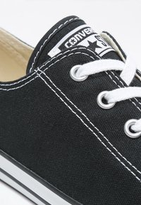 Baskets basses en toile noire avec coutures blanches, lacets blancs, œillets métalliques et étiquette de marque visible sur la languette.