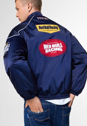 Junge Person trägt eine dunkelblaue Red Bull Racing Jacke mit großen Rückennähten und blaue Jeans, steht mit den Händen in den hinteren Taschen.