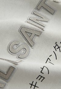 Nahaufnahme eines beigen Stoffs mit aufgestickten großen Buchstaben "SANTA" und schwarzen, gedruckten japanischen Schriftzeichen auf der Oberfläche.