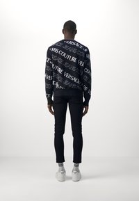 Suéter de punto negro con el texto blanco "VERSACE JEANS COUTURE", combinado con jeans ajustados negros y zapatillas blancas. El modelo está de espaldas.