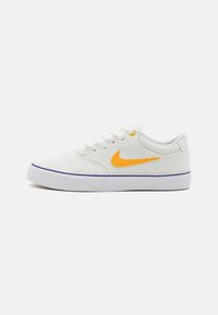 Bílé plátěné tenisky s oranžovým logem Nike, fialovým akcentem na podrážce a tradičním šněrovacím systémem. Plochý, kulatý tvar špičky.