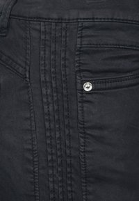 Černá denimová látka s úzkým střihem, vertikálním švem a stříbrným knoflíkem poblíž zadní kapsy.