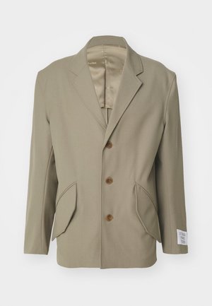Blazer beige avec des revers en pointe, trois boutons marron, deux poches avant et une étiquette sur la manche gauche. Tissu lisse avec un léger brillant.