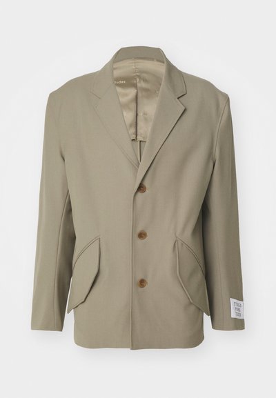 Blazer beige avec des revers en pointe, trois boutons marron, deux poches avant et une étiquette sur la manche gauche. Tissu lisse avec un léger brillant.