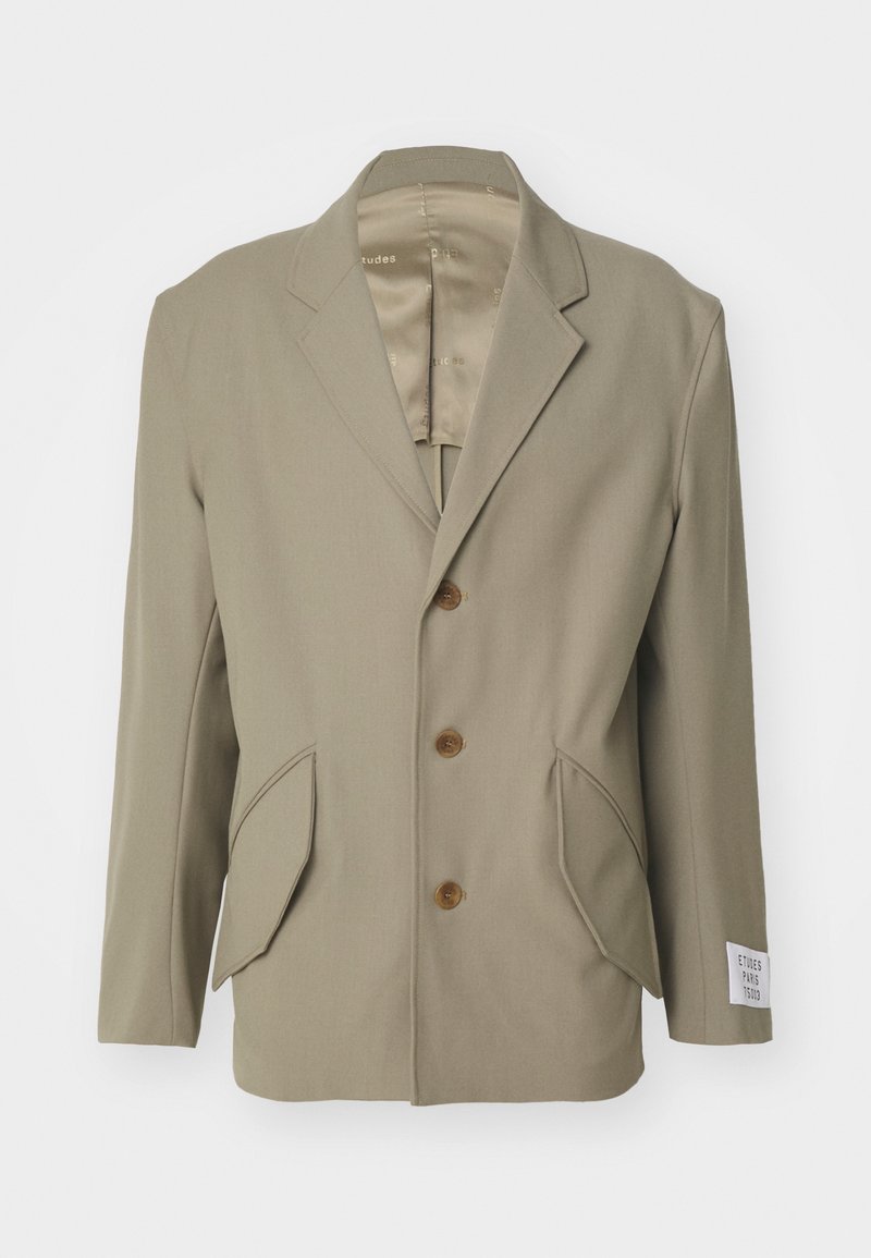 Beige Blazer mit Spitzenrevers, drei braunen Knöpfen, zwei vorderen Taschen und einem Etikett am linken Ärmel. Glattes Material mit leichtem Glanz.