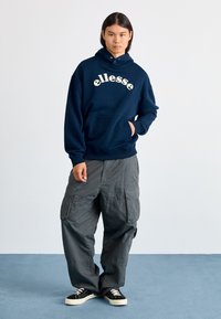 Sudadera marina con el logo de "ellesse" en marfil, con un bolsillo canguro y ribete en el dobladillo. Combinada con pantalones cargo grises holgados y zapatillas negras.