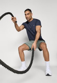 Marine crew-neck t-shirt, grønne shorts, holder et sort kamprep. Hvide atletiske sko, kort hår, fokuseret holdning. Glat, atletisk look.