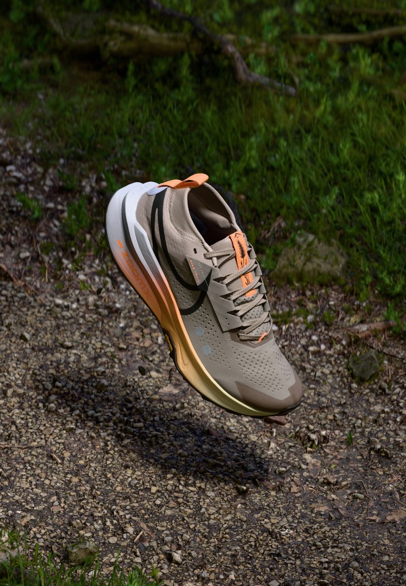 Beige løpesko med mesh-overdel, oransje detaljer, svart swoosh-logo, og en tykk, polstret såle. Teksturerte mønstre på overflaten.