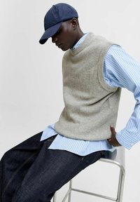 Un pull sans manches en tricot gris porté sur une chemise à boutons rayée bleue et blanche, associé à un jean en denim foncé, assis sur une chaise en métal.