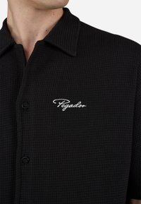 Black textured shirt with a collared neckline, single button, and white embroidered "Pegador" logo on the left chest.
