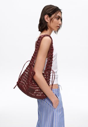 Joven mujer con camiseta sin mangas blanca y pantalones azules a rayas lleva un gran bolso de hombro tejido color granate con detalles de cordón.