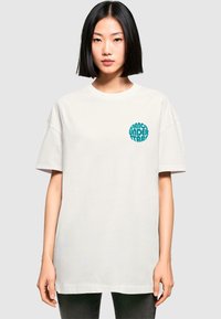 Weißes Baumwoll-T-Shirt im Oversize-Fit mit einem türkisfarbenen, kreisförmigen Grafikdesign, das auf der linken Brustseite "DANCE UNDER STARS" sagt.