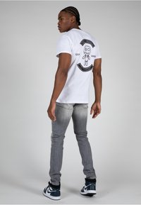 White short-sleeve t-shirt with a black printed graphic on the back. Gray slim-fit jeans and black and teal sneakers complete the outfit.