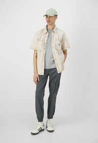 Chemise à boutons beige à manches courtes au-dessus d'un t-shirt gris, associée à un pantalon gris foncé et des baskets blanches avec des accents noirs. Casquette vert clair.