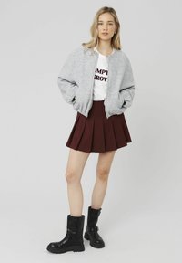 Veste bomber grise, t-shirt blanc avec texte rouge, jupe plissée bordeaux et bottines noires à détail de boucle. Textures lisses, design décontracté.