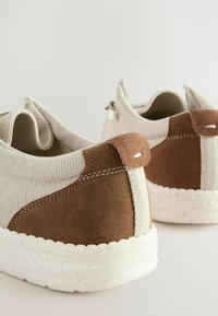 Kangasneakers dengan bagian atas beige dan aksen suede coklat serta sol karet putih bertekstur. Dilengkapi dengan tab tarik belakang untuk memudahkan pemakaian.
