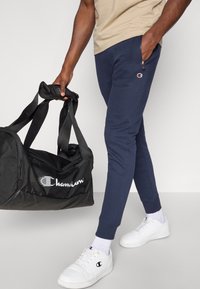 Bolsa de lona negra con un logo blanco de "Champion", llevada por una persona con pantalones deportivos azul marino y zapatillas blancas con detalles visibles de la marca y cremallera.