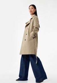 Trench beige con botones grandes, bolsillos laterales y cintura ceñida con cinturón, combinado con jeans oscuros de pierna ancha y zapatos negros de punta.