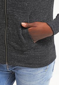 Mörkgrå zip-up hoodie i strukturerad tyg. Har en framficka och åtsittande mudd, kombinerad med ljusblå denimjeans.