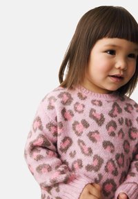 Kleinkind mit glattem braunem Haar, das einen rosa und braunen Leopardenmuster-Pullover trägt und leicht zur Seite blickt, mit neutralem Gesichtsausdruck.