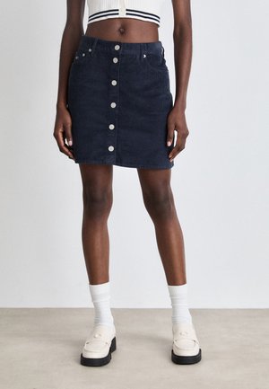 Woman wearing a navy blue button-front corduroy mini skirt, white cropped top, white socks, and cream loafers standing on a gray floor.