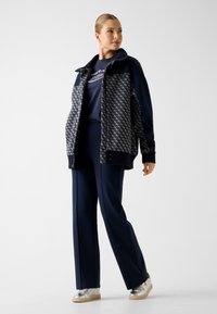 Marineblauw en patroonrijke ritsjas met een kraag, gecombineerd met een donkerblauw T-shirt en bijpassende op maat gemaakte broek. Zilveren sneakers maken de outfit compleet.