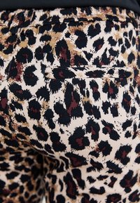 Pantalon à motif léopard en beige, noir et bordeaux. Présente une poche arrière et un tissu extensible à la texture lisse. Ceinture ajustable.